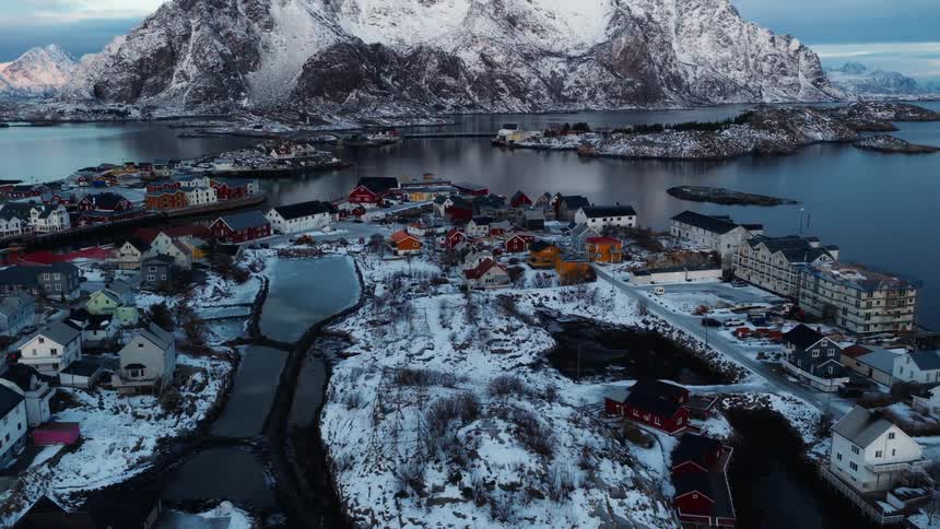 An aerial drone shot pulls back from a colorful fishing village to reveal the breathtaking, majestic, snow-covered mountains of the Lofoten Islands in Norway