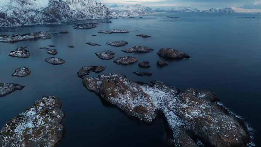 An aerial drone shot pans up over snow-dusted islands to reveal the epic, wintry mountain landscape of a Norwegian fjord at twilight