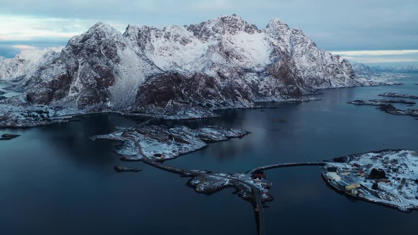A breathtaking aerial view slowly pulls back to reveal the magnificent winter landscape of the Lofoten Islands in Norway, with snow-covered mountains towering over the sea and interconnected islands