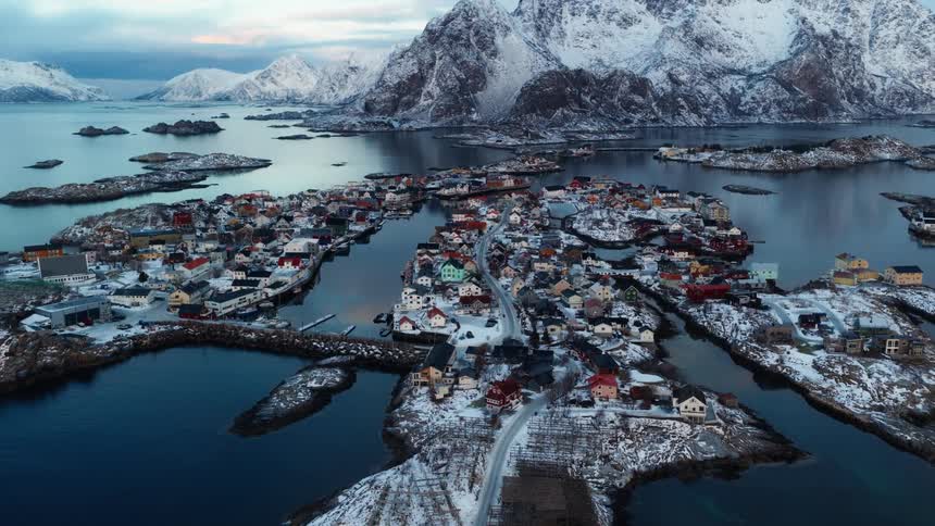 A stunning aerial shot pulls back to reveal a quaint fishing village scattered across a snowy archipelago, nestled between calm waters and majestic mountains