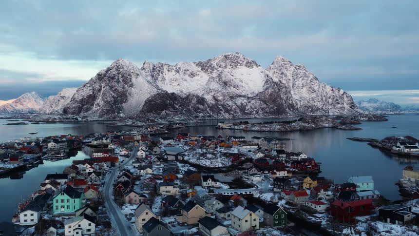 A stunning aerial pull-back shot reveals a picturesque, snow-covered coastal village with towering mountains reflected in the calm waters