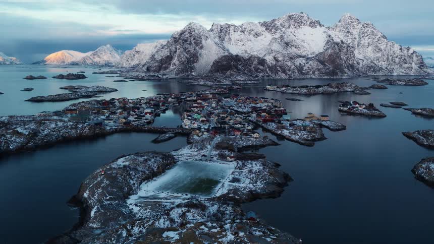 A breathtaking aerial shot captures a snowy island village and a football field in Norway, set against majestic mountains as the sun rises
