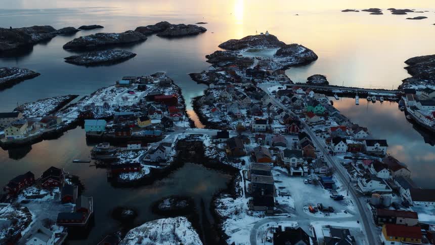 A stunning aerial shot tilts up from a snowy coastal town on an archipelago to reveal a beautiful golden sunset over the tranquil ocean