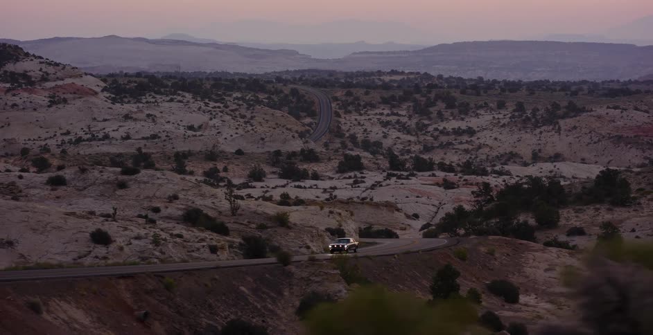 A classic car with its headlights on drives along a winding scenic byway through a vast, rocky desert landscape at dusk