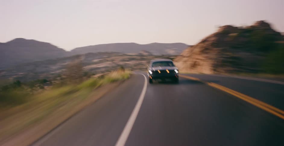 A classic black muscle car speeds down a scenic and winding desert road at twilight