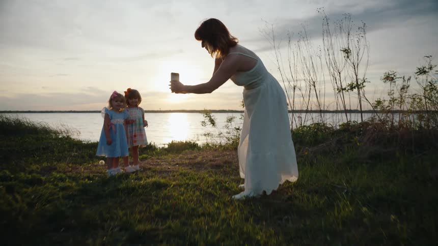 Slow dolly shot of a mother taking a smartphone picture of her two young daughters posing together by a lake at sunset