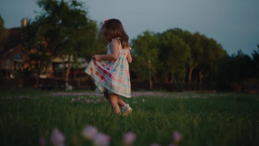 A cute little girl in a colorful dress joyfully runs and twirls through a grassy field of wildflowers at dusk in slow motion
