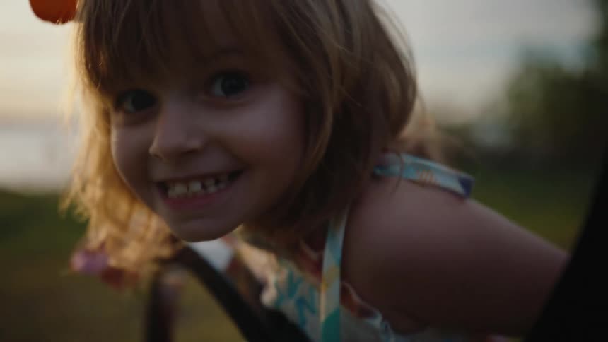 A cute little girl with a big orange bow in her hair smiles happily at the camera while sitting on a park bench at sunset