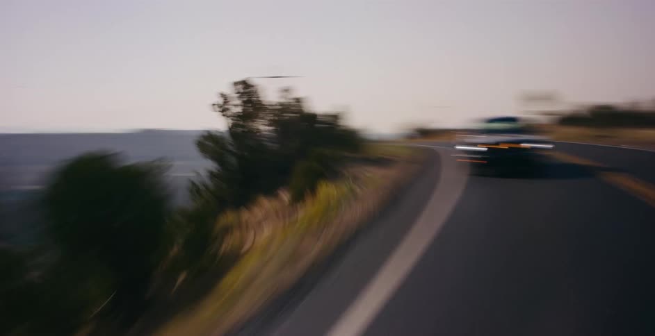 A classic black Ford Mustang cruises along a winding scenic road through a mountainous desert landscape at dusk