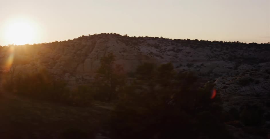 Drone footage pulls back over a vast desert canyon landscape as the sun sets, creating a golden lens flare