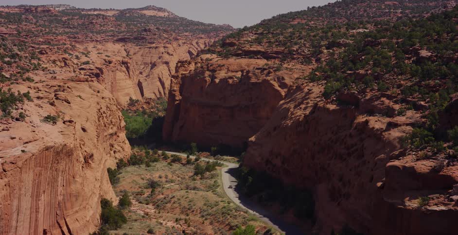 A dramatic sliding shot reveals a lone car driving on a winding road through a majestic red rock canyon