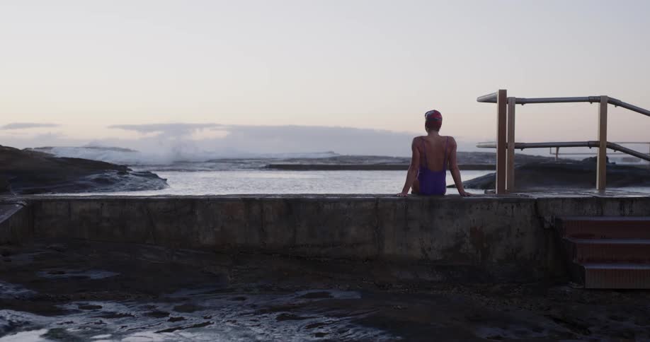 A female swimmer in a purple swimsuit sits on the edge of a rocky ocean pool at dawn, watching the waves crash in the distance