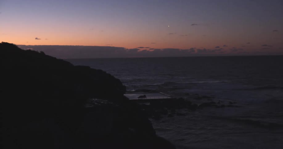 A silhouetted headland overlooks a calm ocean and a rock pool as twilight fades in the sky