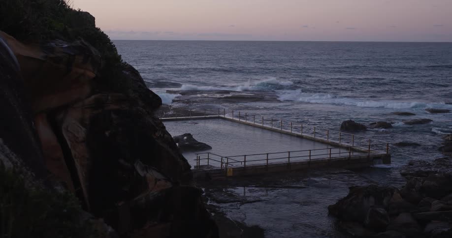 A lone swimmer glides through the calm water of a coastal rock pool in slow motion as gentle waves break against the shore at dawn