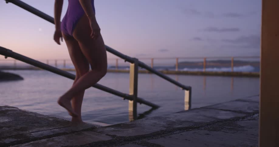 A female swimmer in a purple swimsuit and red cap walks along the edge of an ocean pool and slowly enters the water at sunrise