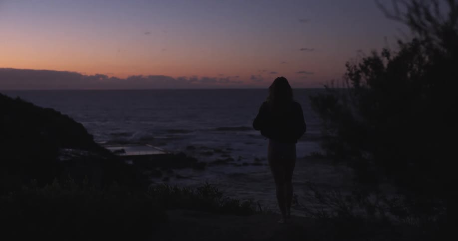 A silhouetted woman walks to a viewpoint to watch the ocean waves crash against the rocky shore at twilight