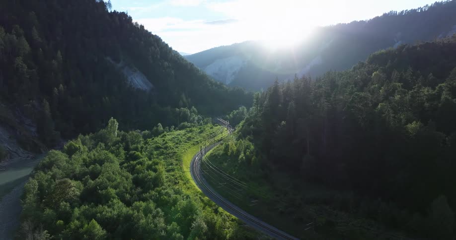 An aerial view reveals a scenic railway winding through a lush, sunlit mountain valley next to a river