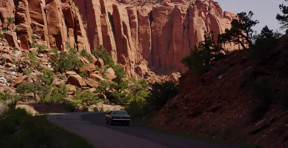 A classic green muscle car drives on a winding road through a majestic red rock canyon in the desert