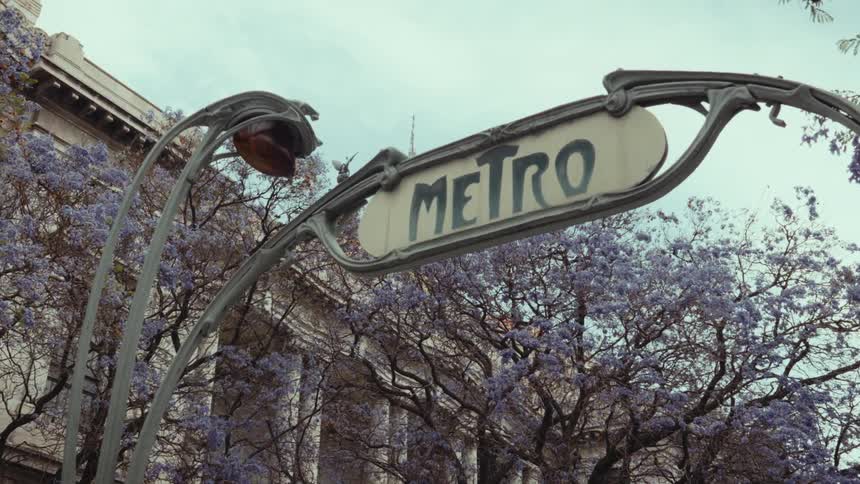 A classic Art Nouveau metro sign is framed by blooming purple Jacaranda trees in a historic city center