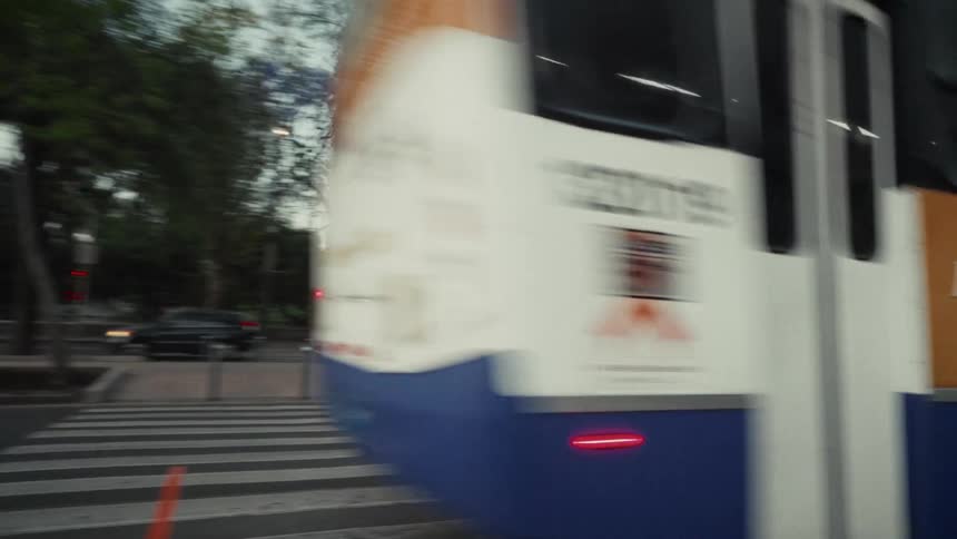 A blue city bus drives past the camera through a crosswalk on a tree-lined street at dusk
