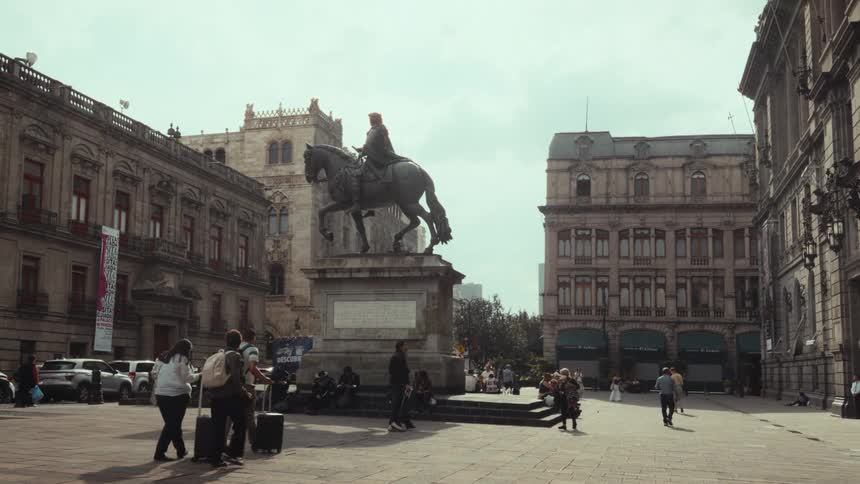 A wide, static shot of the historic Plaza Manuel Tolsá in Mexico City, where people walk and relax around the iconic equestrian statue of Charles IV