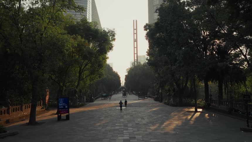 Two people walk down a wide, tree-lined promenade in a city park as the golden hour sun rises in the distance
