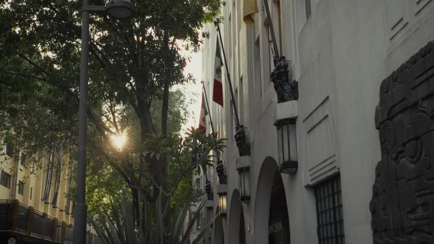Sunlight filters through the trees, illuminating an elegant building with the Mexican flag and pre-Hispanic carvings in Mexico City