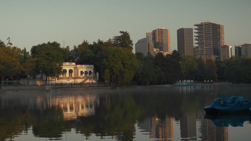 A tranquil sunrise casts a golden glow over a misty urban park lake, perfectly reflecting a historic building and a modern city skyline