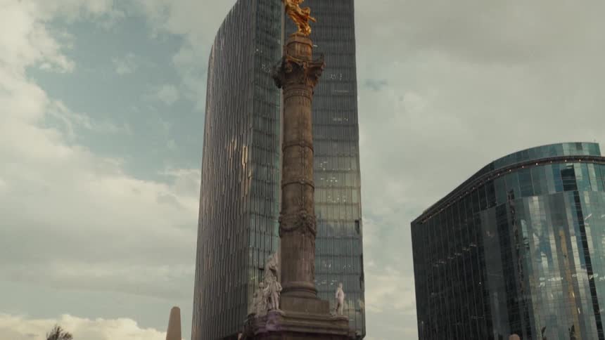 A slow tilt up reveals the iconic Angel of Independence monument set against modern skyscrapers in Mexico City on a cloudy day