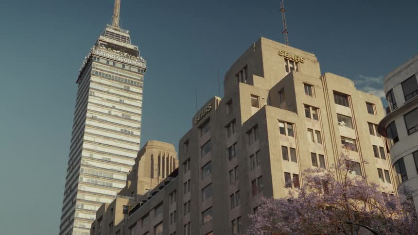A stately low-angle view pans up to reveal the iconic Torre Latinoamericana skyscraper in Mexico City, framed by vintage buildings and purple jacaranda trees