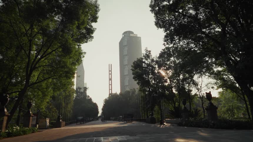 The sun rises over the modern skyline of Mexico City, viewed from a tranquil, tree-lined park walkway in the early morning haze