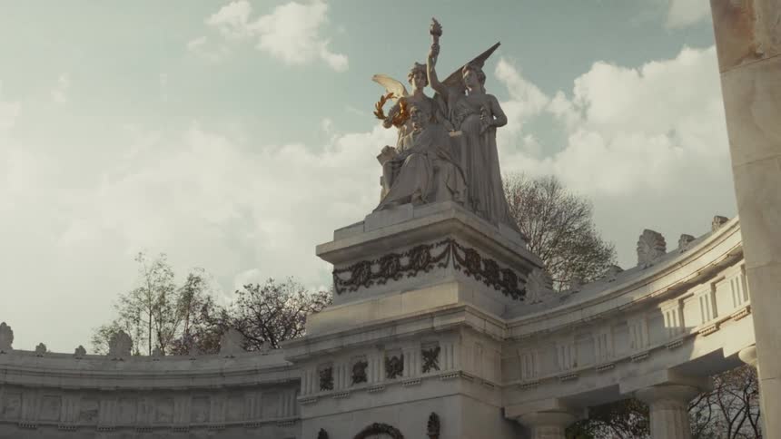 A low-angle tracking shot of the historic Hemiciclo a Juárez monument in Mexico City, showcasing its grand marble statues and columns