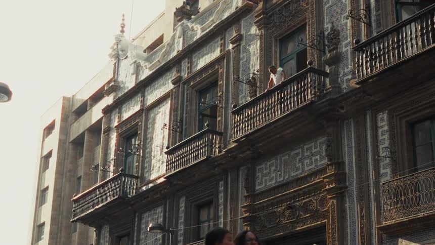 A man on a balcony watches pedestrians pass by an ornate historic building on a sunny day