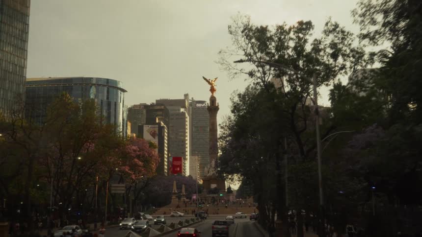 A slow-motion cinematic drive down a tree-lined avenue towards the Angel of Independence in Mexico City at dusk