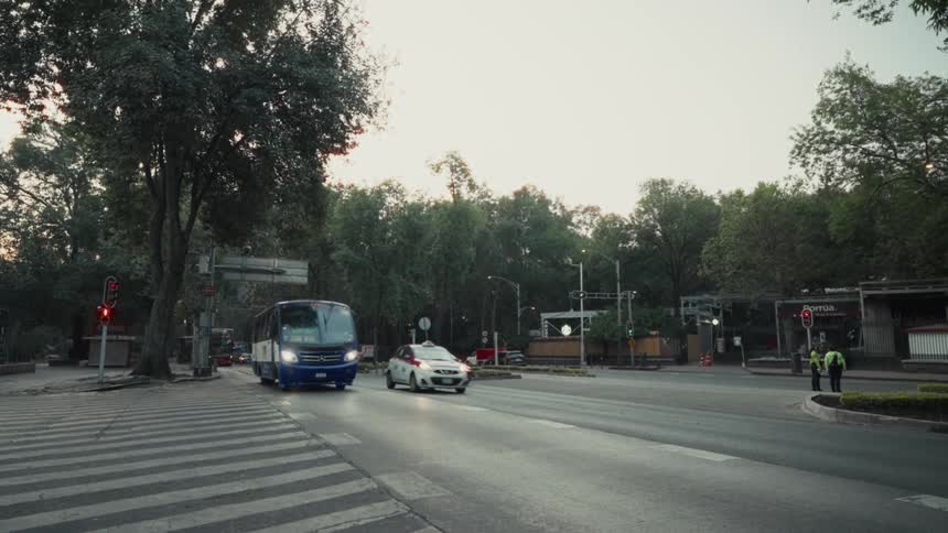 A CDMX bus navigates through evening traffic on a tree-lined street in Mexico City as the camera pans to follow its movement