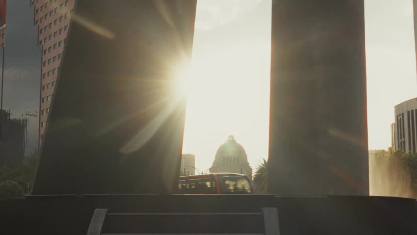 The sun flares behind the Monument to the Revolution in Mexico City, framed by a modern monument as a red tour bus drives past