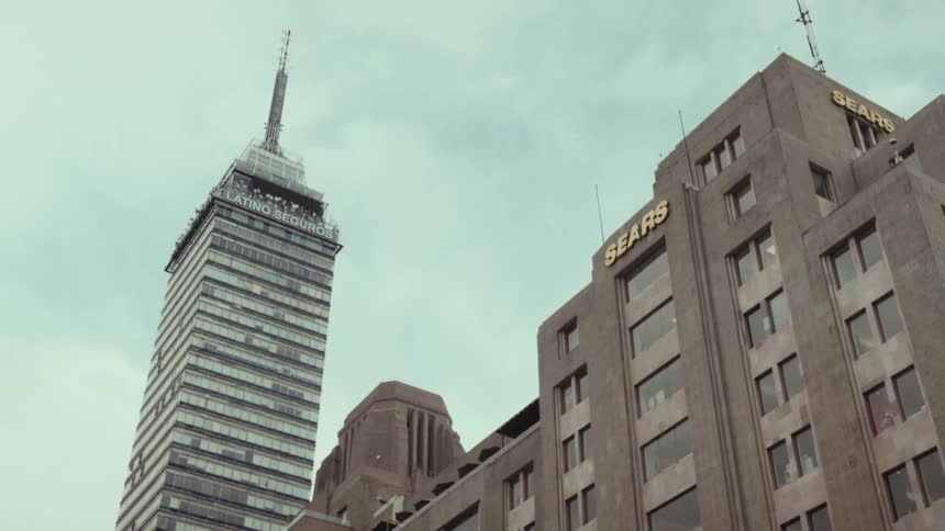 A low-angle view shows the iconic Torre Latinoamericana skyscraper next to a Sears department store in Mexico City under a cloudy sky