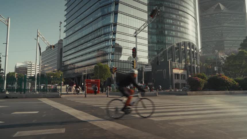 A low-angle view of a city street shows cyclists riding across a crosswalk in front of modern glass skyscrapers