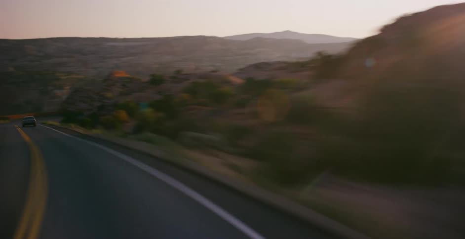 A time-lapse point-of-view drive follows a vintage car on a winding highway through a desert canyon landscape at sunset