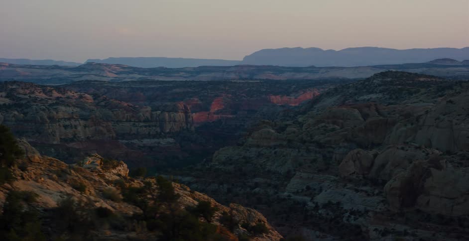 An aerial view pans over a vast, rocky desert canyon landscape with red rock formations glowing at dusk
