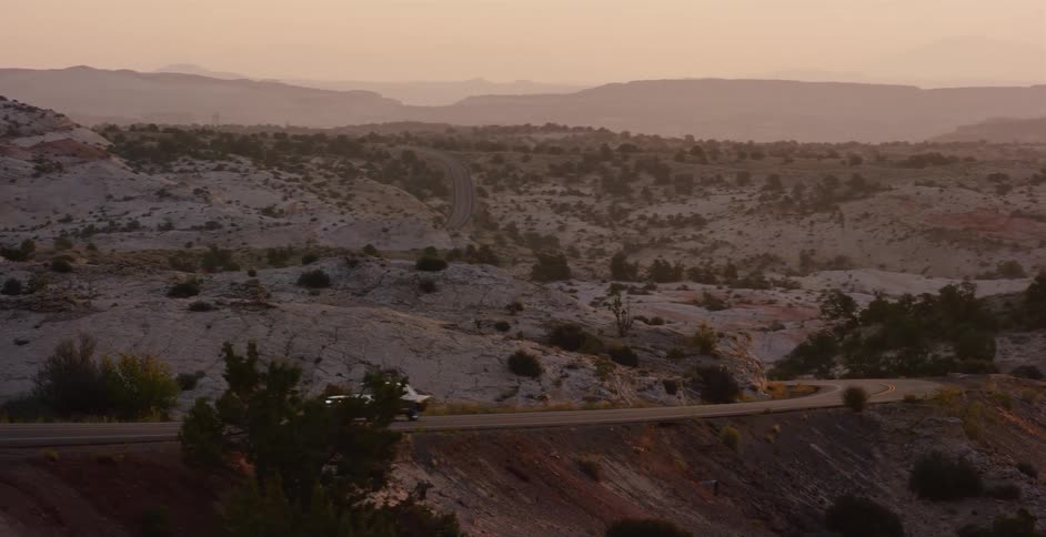A classic car cruises along a winding road through a vast, rocky desert landscape during a golden sunrise