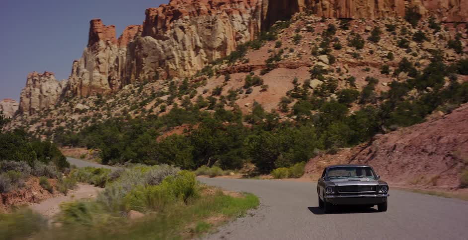A classic grey Ford Mustang drives along a winding road through a majestic red rock canyon in the desert