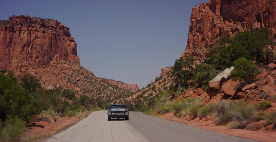 A classic car drives away down a remote road through a majestic red rock canyon