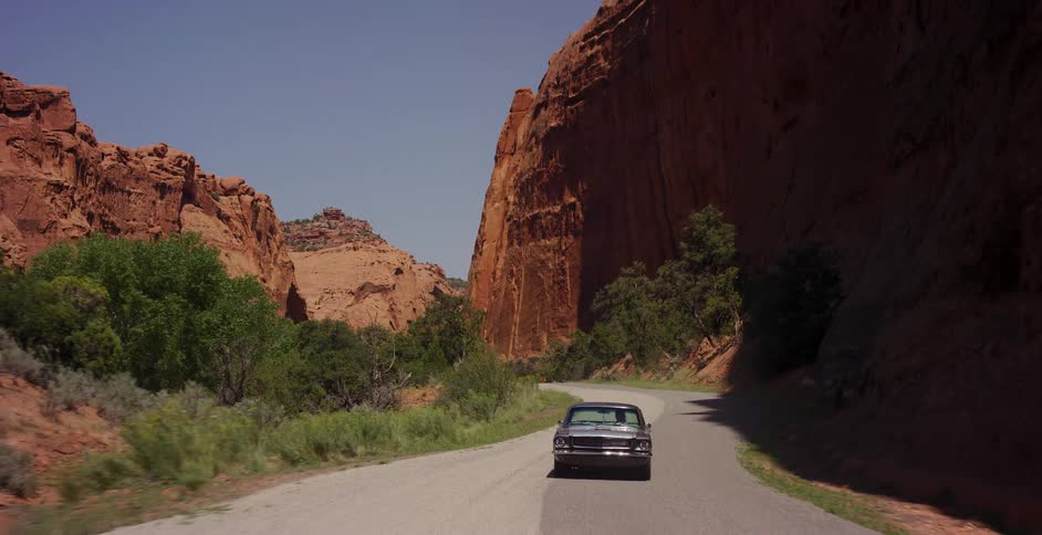 A classic American muscle car drives down a winding road through a majestic red rock canyon on a sunny day