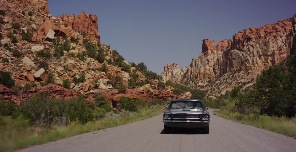 A classic silver muscle car drives steadily down a scenic highway through a magnificent red rock desert canyon