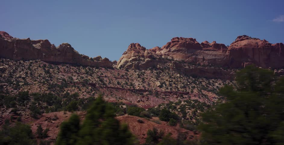 A slow-moving drive-by shot showcases the layered, multicolored rock formations of a vast desert mountain range under a clear blue sky