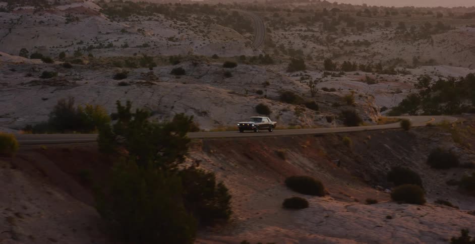 A vintage black muscle car drives along a winding scenic highway through a desert landscape at sunrise