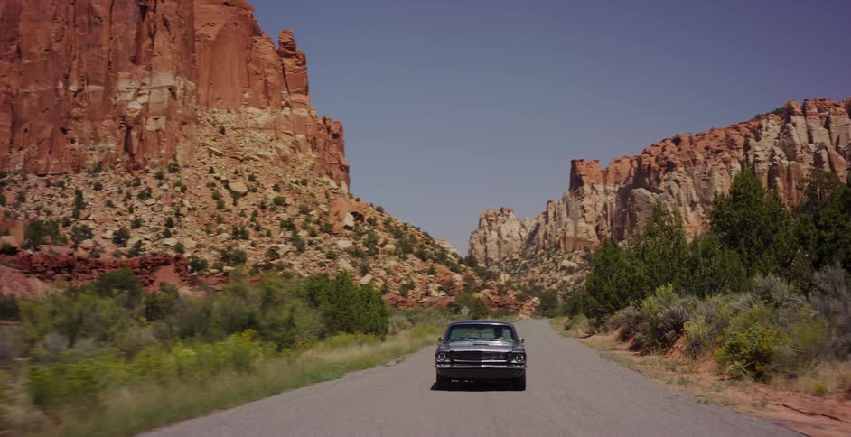 A classic muscle car cruises down a scenic highway through a vast red rock canyon on a sunny day