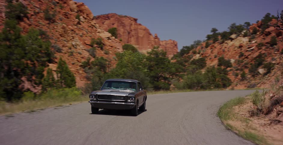 A classic silver muscle car cruises down a scenic desert highway surrounded by red rock canyons