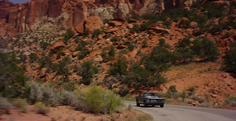 A classic muscle car drives along a scenic, winding road through a dramatic red rock canyon during the day